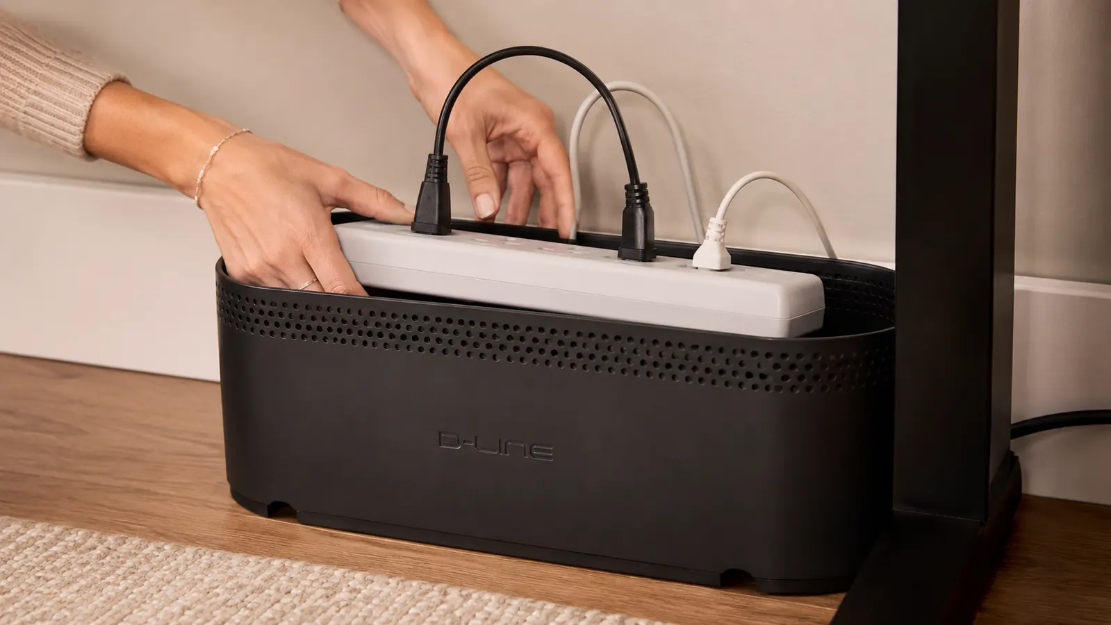 Hand tucking a bundle of cords and a power strip into an open cream cable management box under a desk