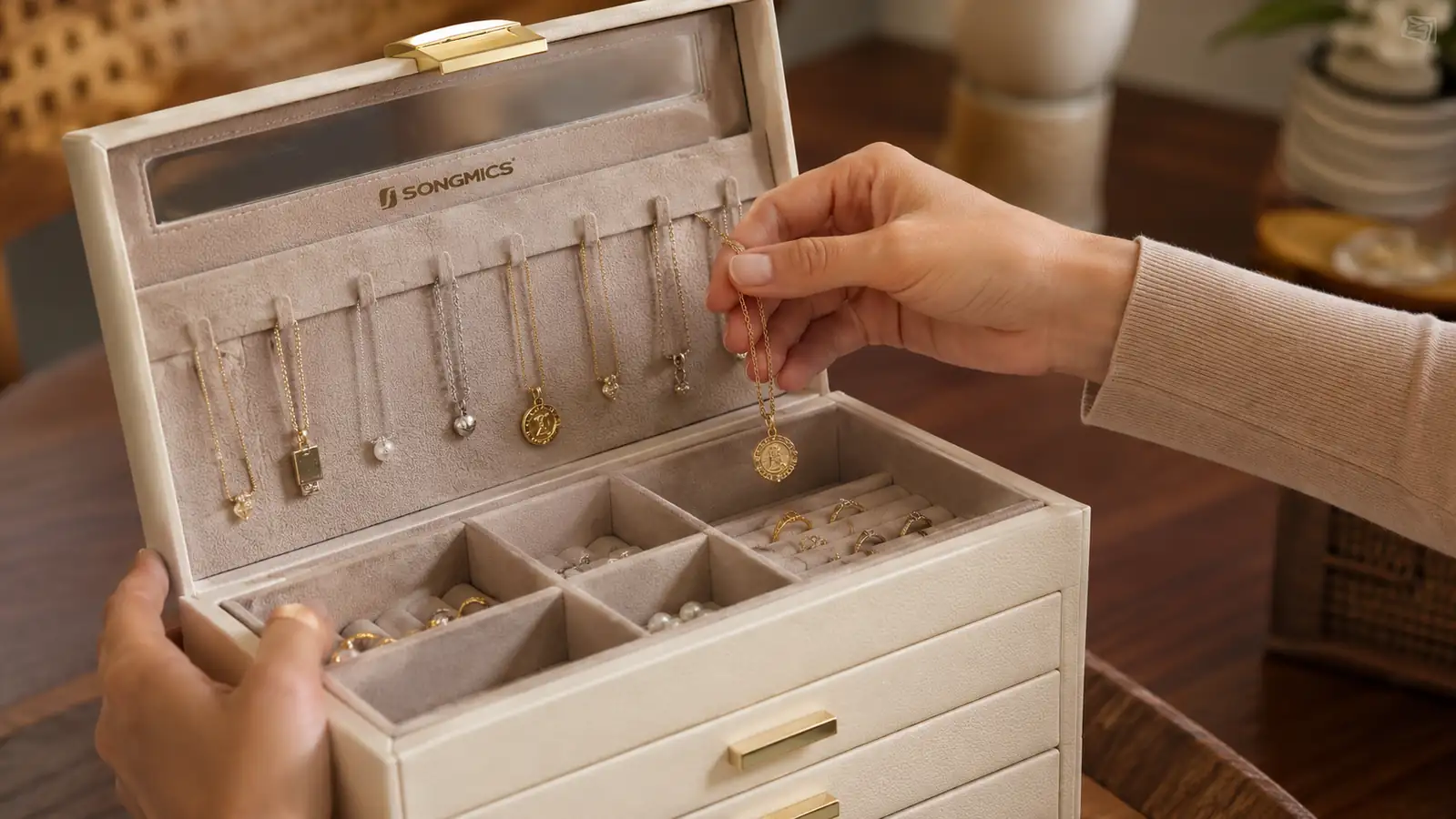 Woman's hand lifting a necklace off an interior hook inside an open glass-lid jewelry box