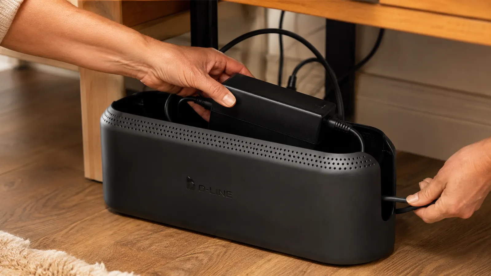 Hand feeding a laptop dock power brick and cords through the cutout of an open D-Line cable box under a desk